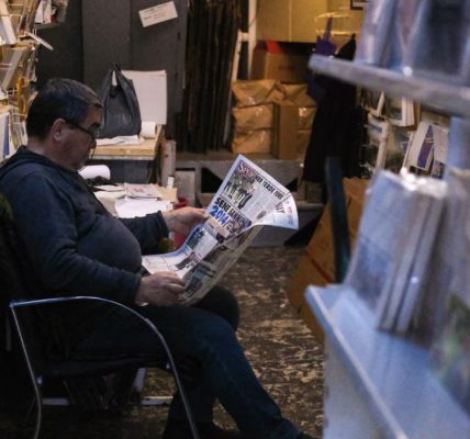 Gentleman sits comfortably reading a newspaper in a cozy, dimly lit bookstore.